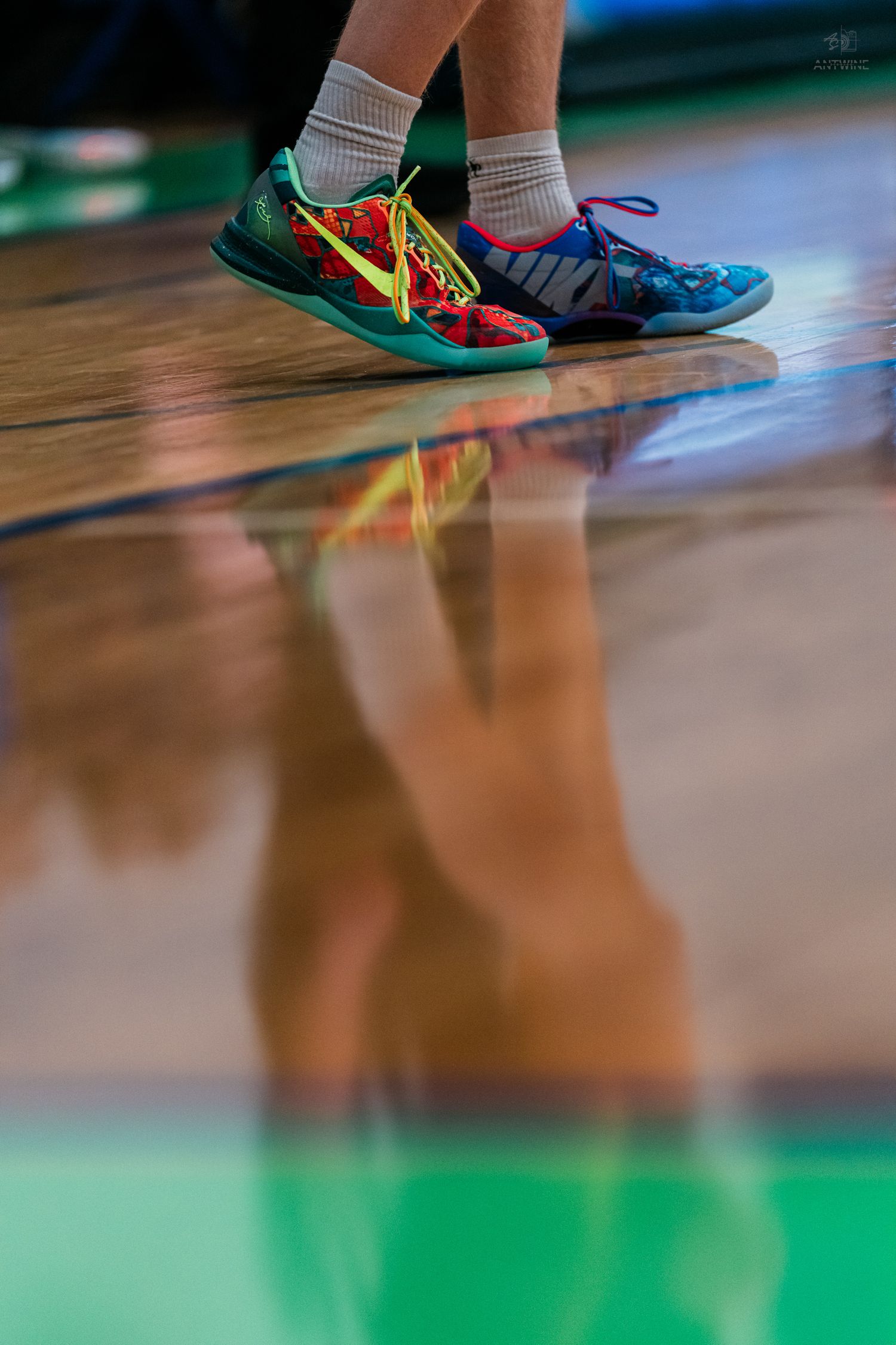 A basketball player wearing colorful Nike sneakers with yellow laces reflected on a shiny hardwood floor.