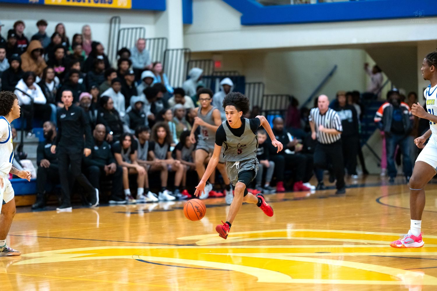 Basketball player in gray uniform dribbles aggressively down the court during a high school gymnasium game.