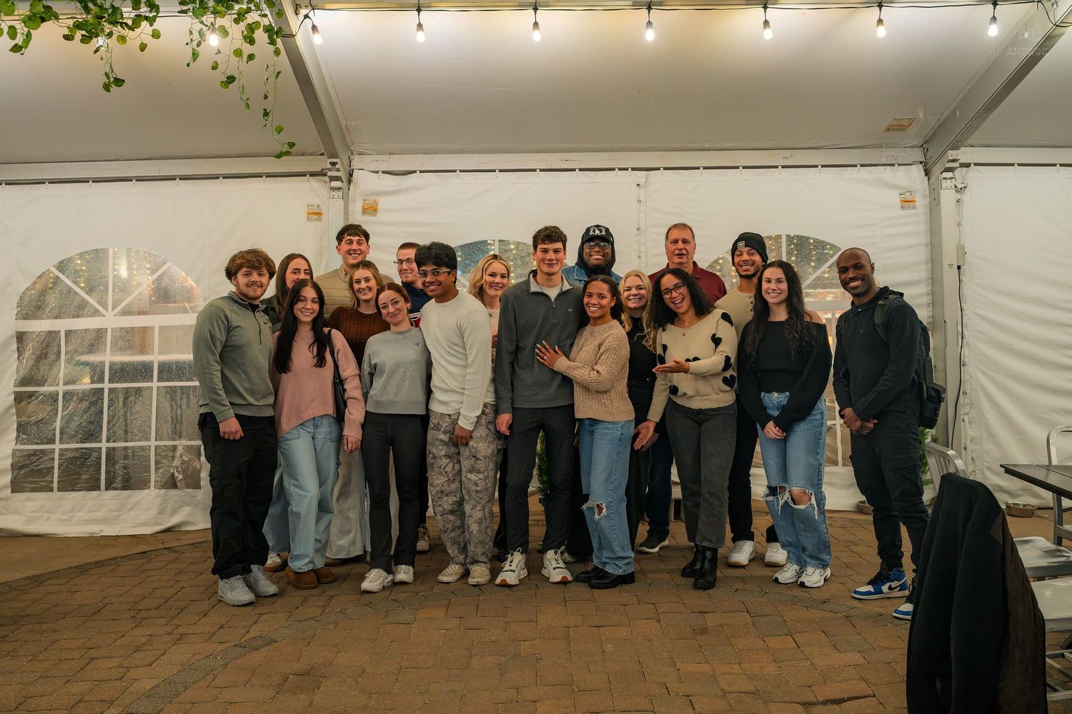 Large group of young adults smile together under string lights in white tent.