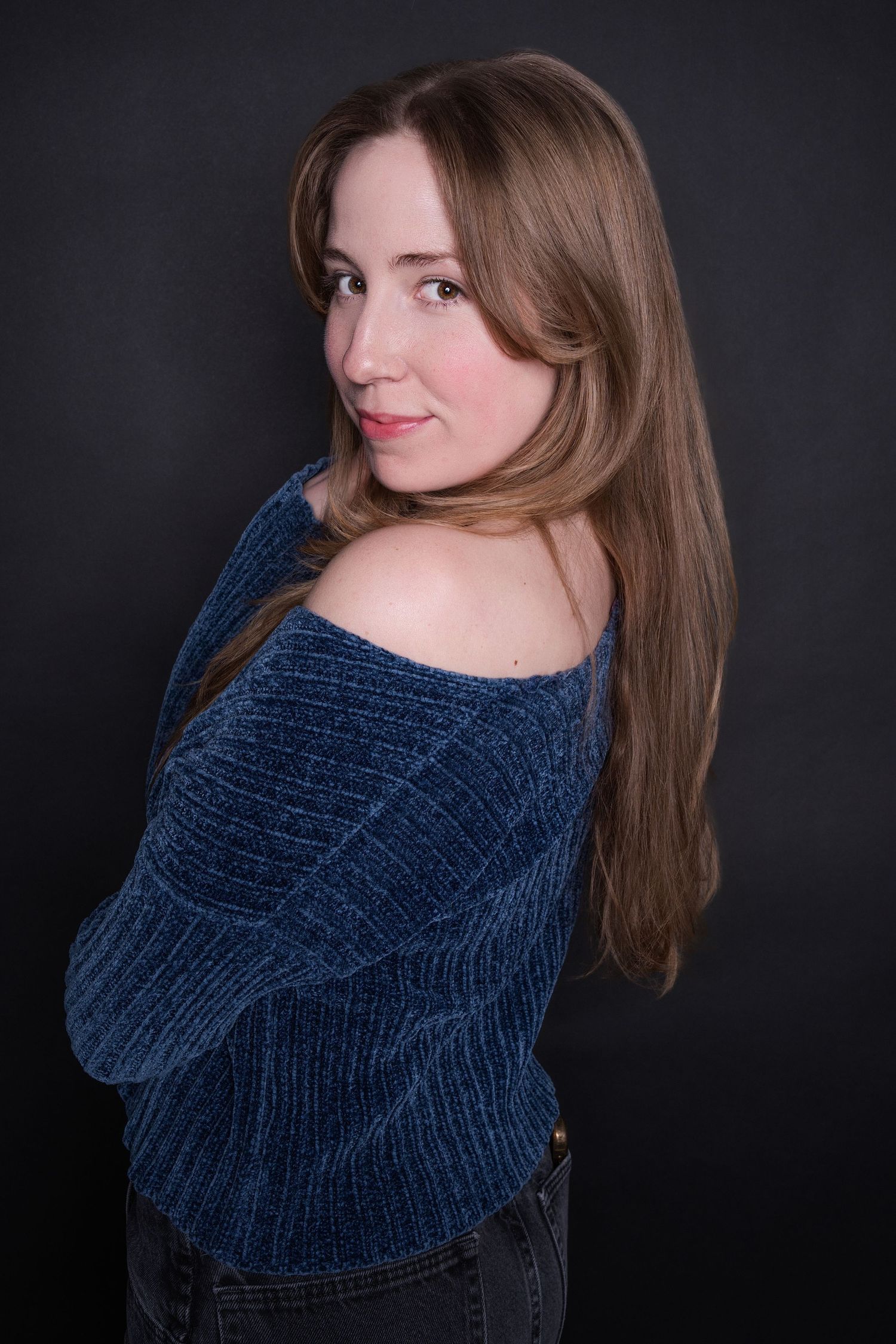 Woman in blue off-shoulder sweater posing sideways against dark background.