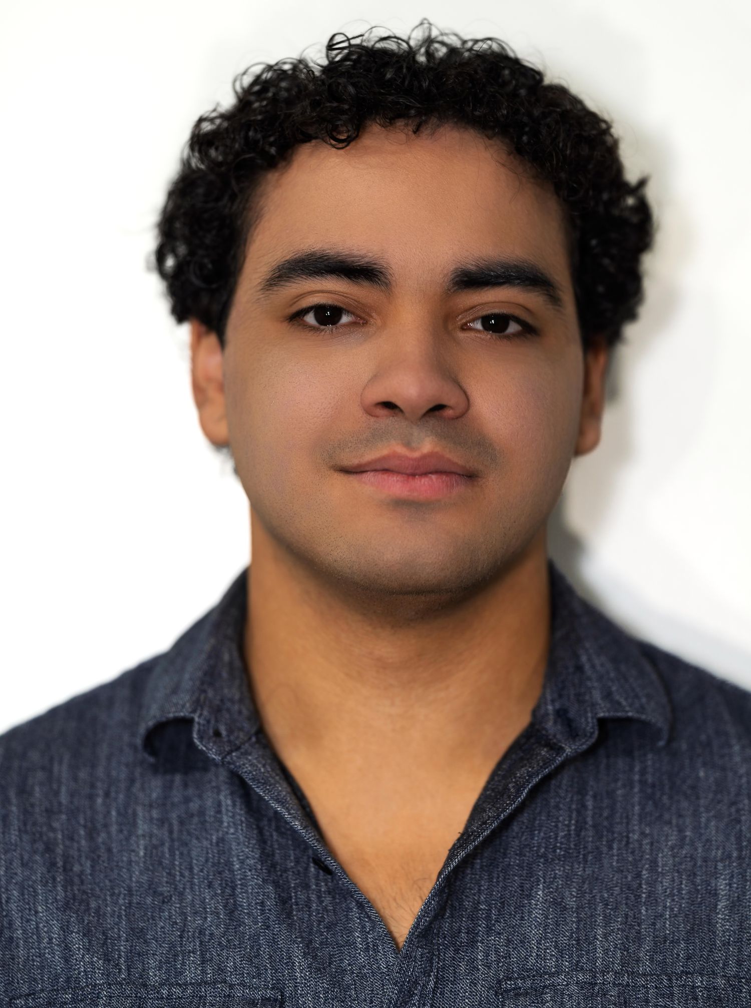 A young man with curly dark hair in a gray collared shirt poses with a neutral expression in a portrait.