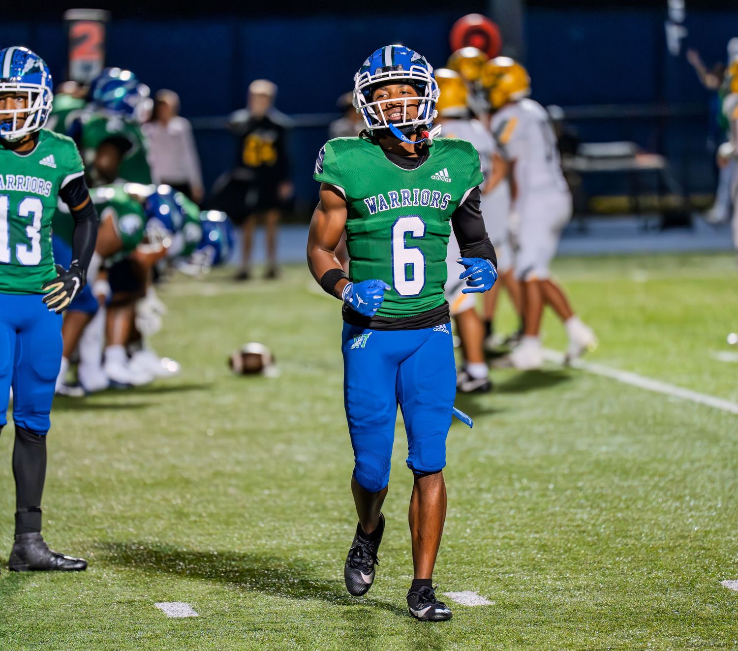 High school football player wearing number 6 in green Warriors jersey walks on field during night game.