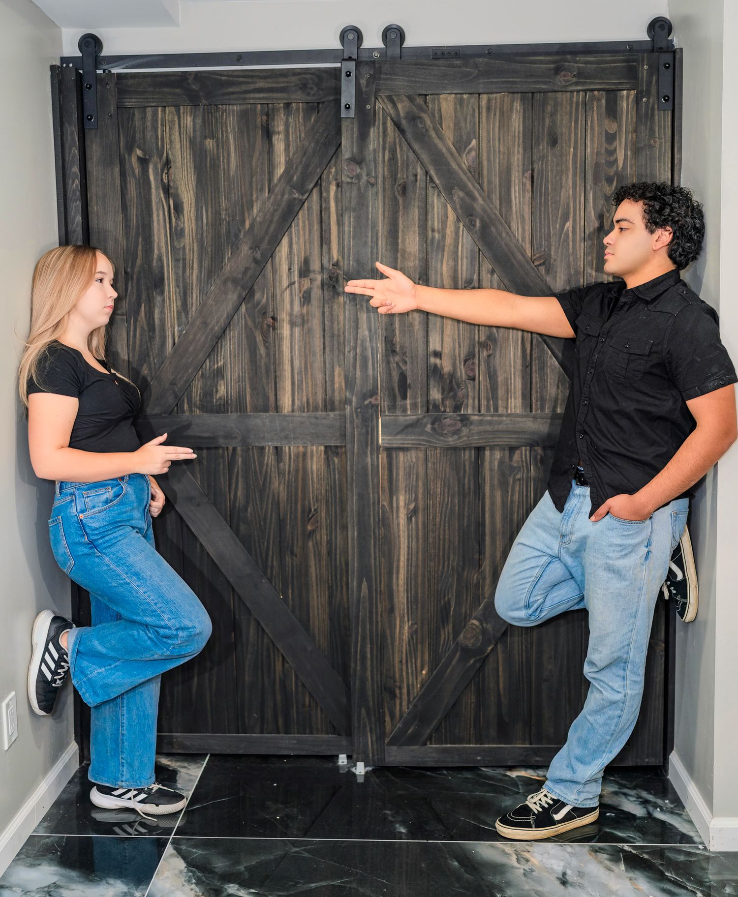 Young couple standing apart by dark wooden barn door, man pointing toward woman.