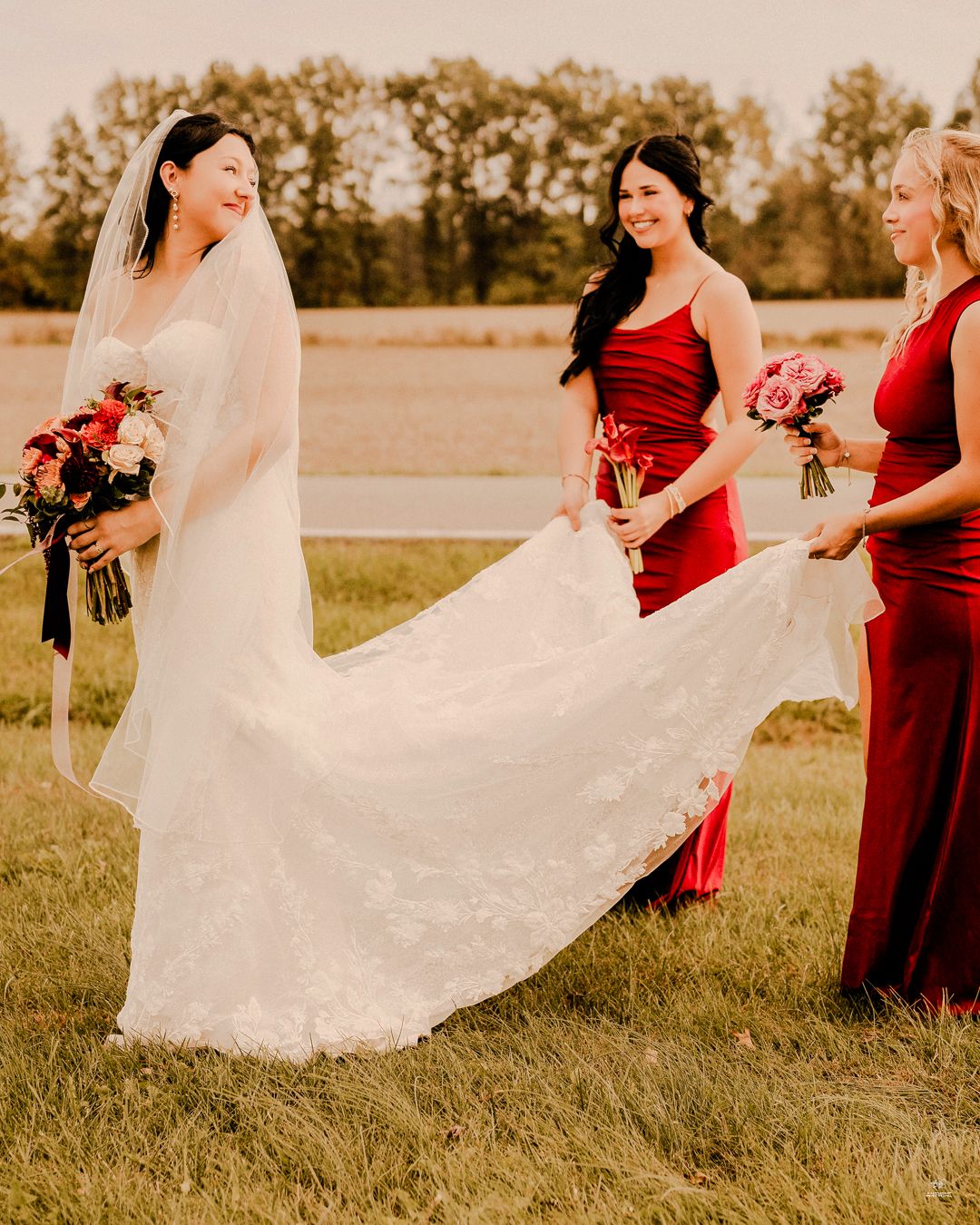 Smiling bride in lace wedding gown with veil, flanked by two bridesmaids in red dresses holding bouquets.