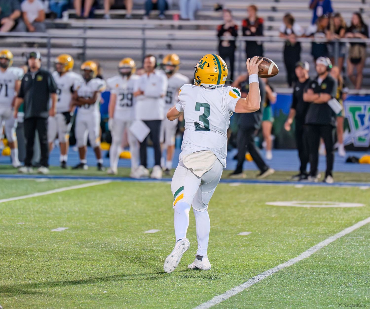 High school football player wearing number 3 green and gold uniform throws a pass during a Friday night game.