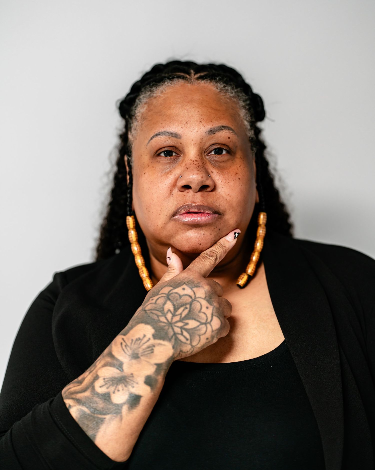 Woman with braided hair and floral hand tattoo poses thoughtfully, wearing gold earrings and black outfit.