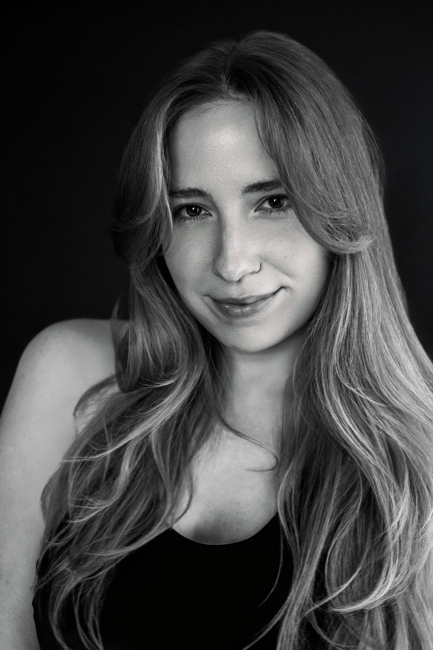 Black and white close-up portrait of young woman with long wavy hair and nose piercing against dark background.