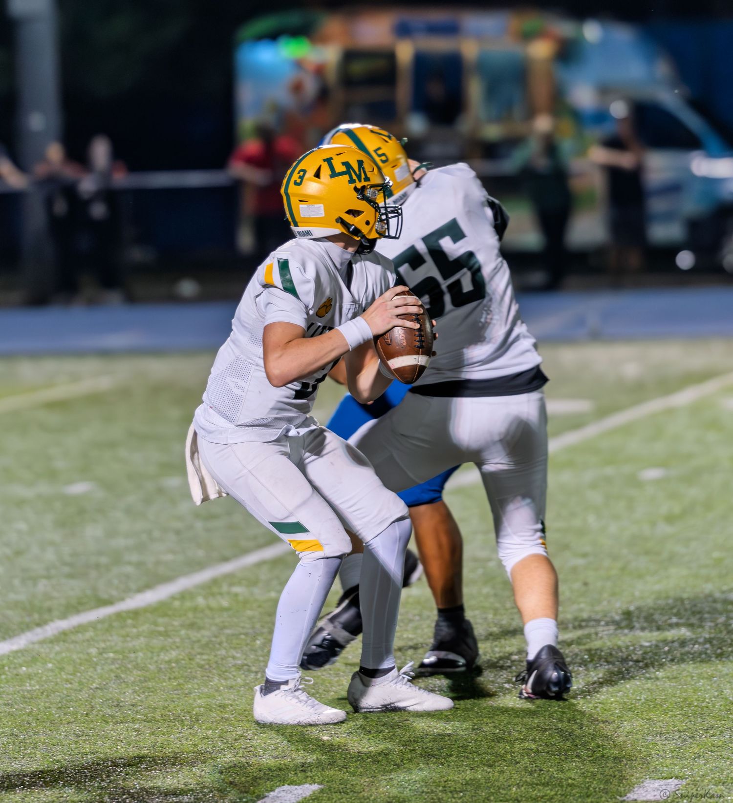 High school football quarterback receives snap from center #65 during a night game.