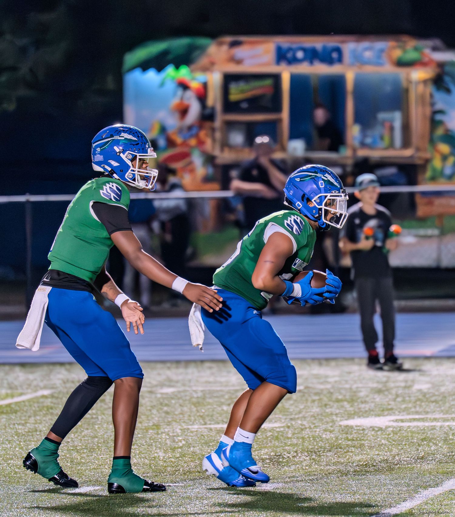 A quarterback in green and blue uniform hands off the football to a running back during a night game.