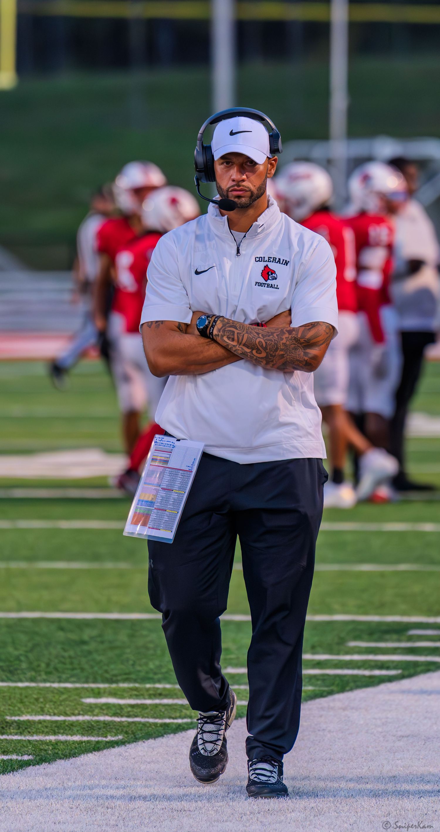 Colerain Football coach in white Nike polo and cap walks the sideline during a high school football game.
