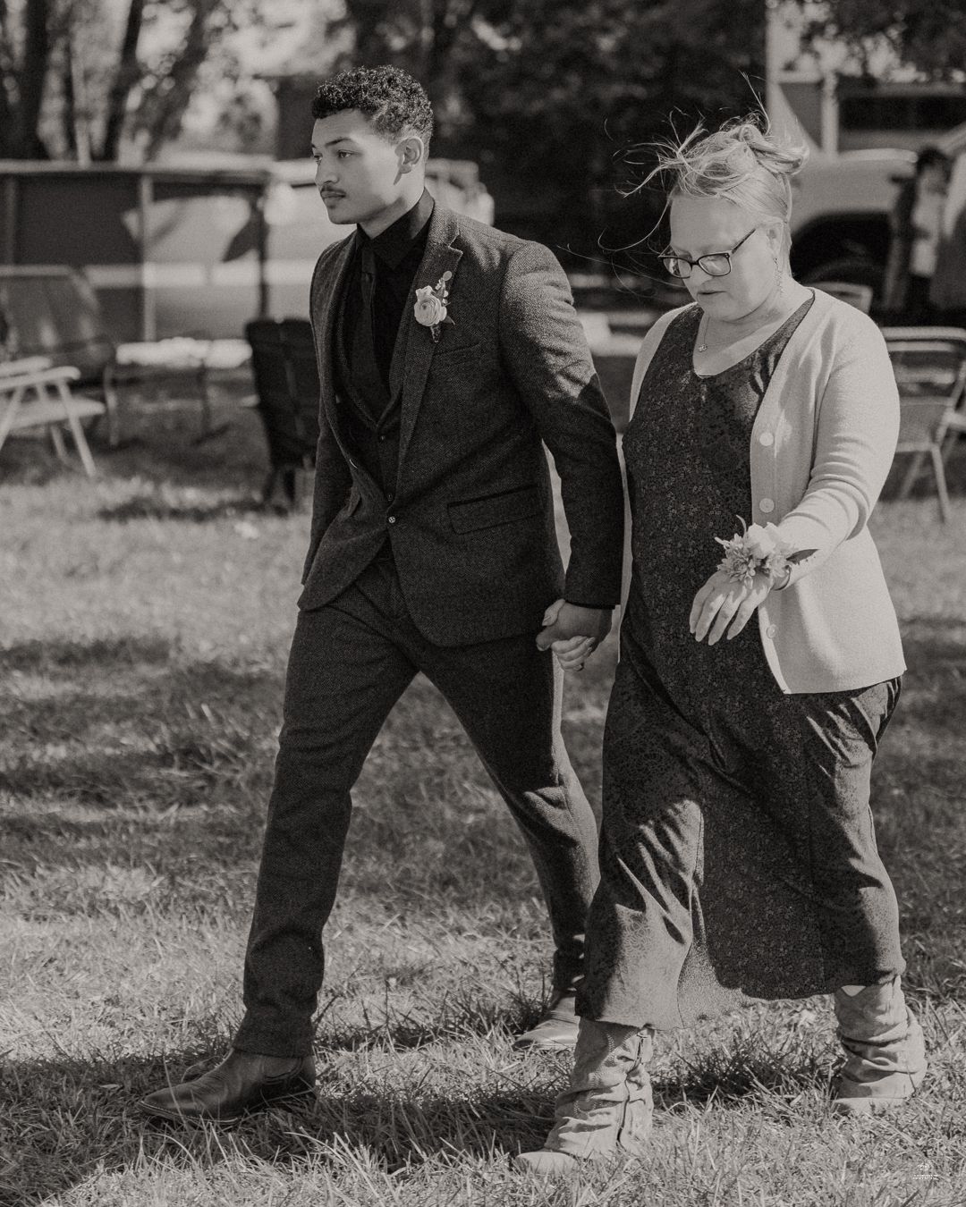 Black and white photo of groom in suit with boutonniere escorting woman at outdoor wedding.