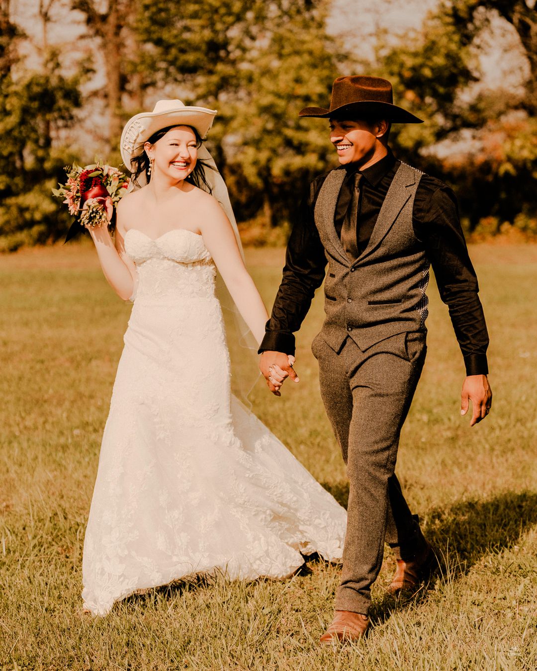 Bride and groom in cowboy hats walk hand in hand through a sunny green field on their wedding day.