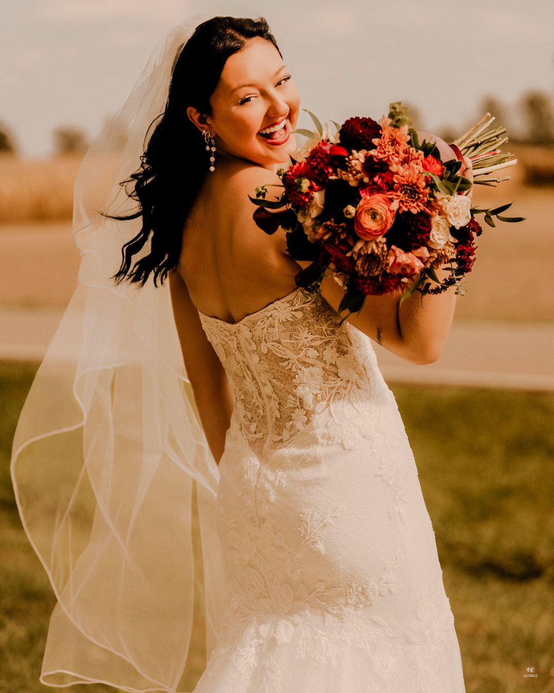 A smiling bride in a strapless lace gown holds a vibrant red and orange bouquet in golden hour light.