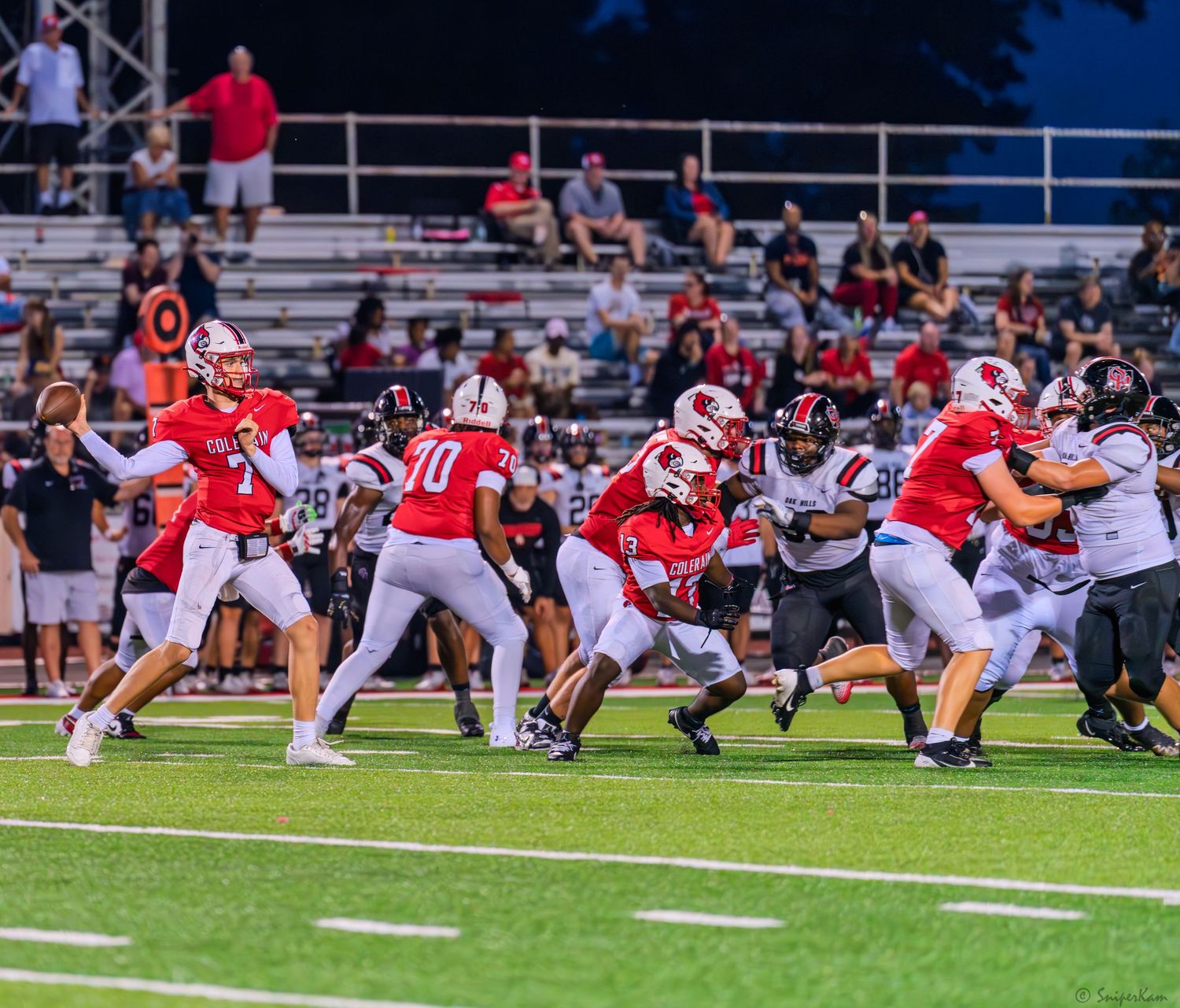 High school football players in red uniforms execute a play during a night game with fans in the stands.