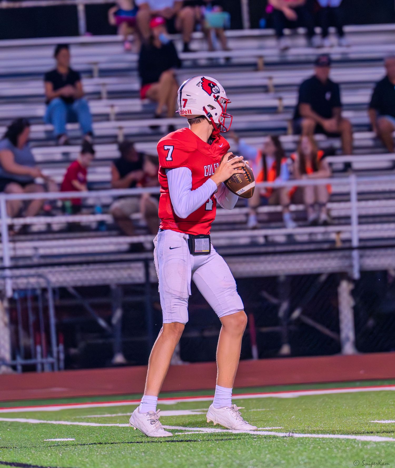 High school quarterback number 7 in red uniform prepares to throw during a night football game.