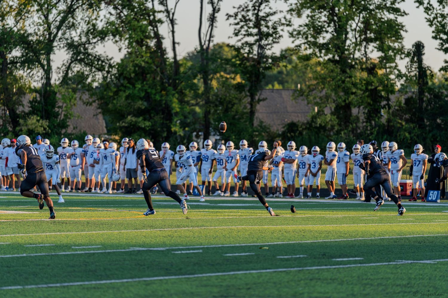Football players run on green field during evening high school game.