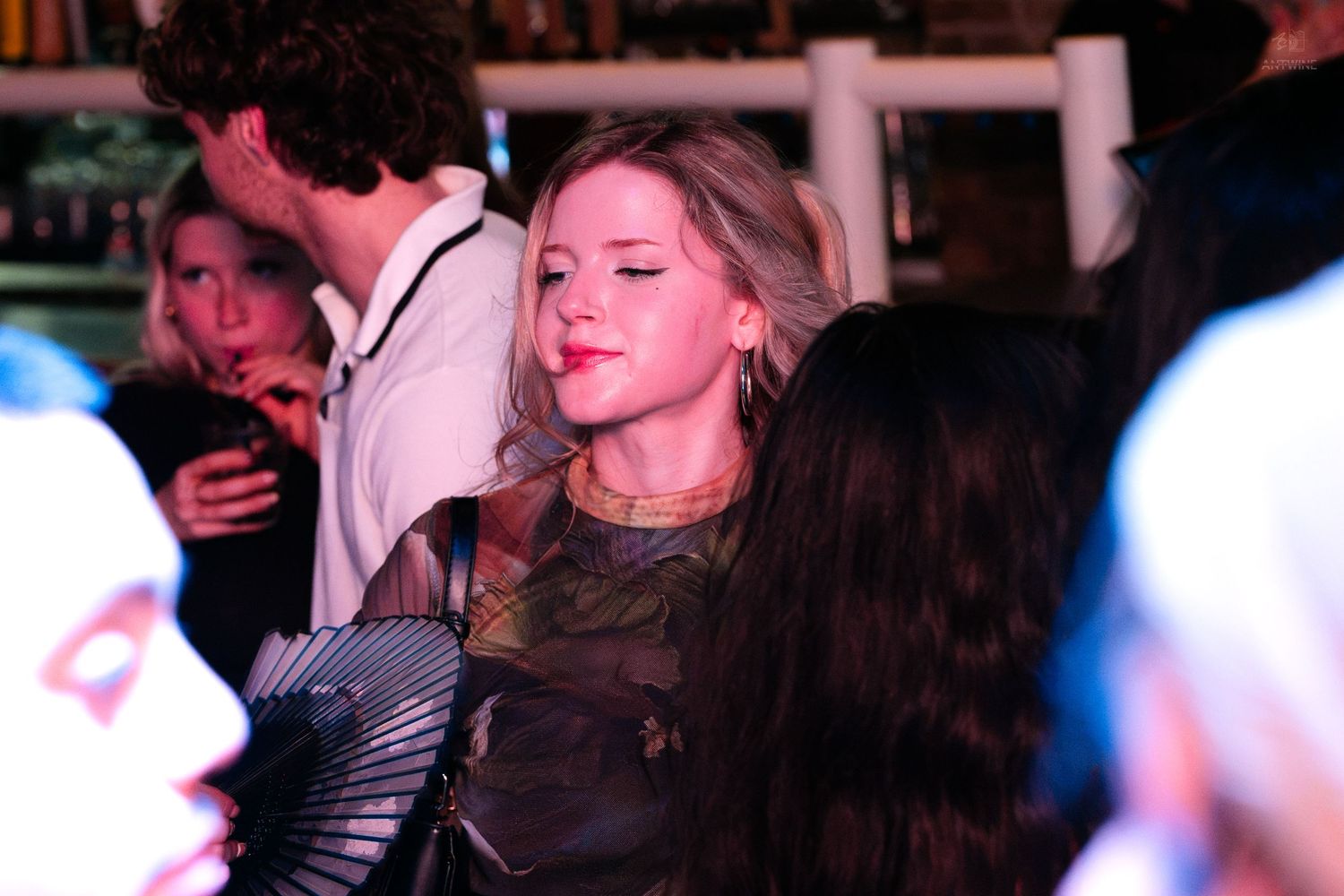 Blonde woman holding a decorative fan at a busy bar or nightclub event.