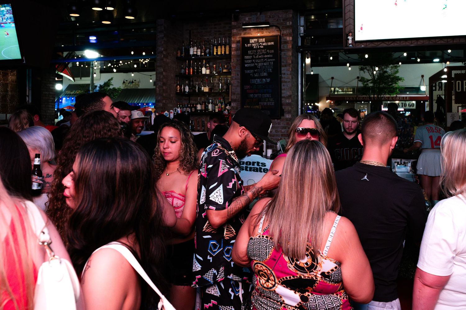 Crowd of people socializing at a busy bar with TVs and shelves of bottles.