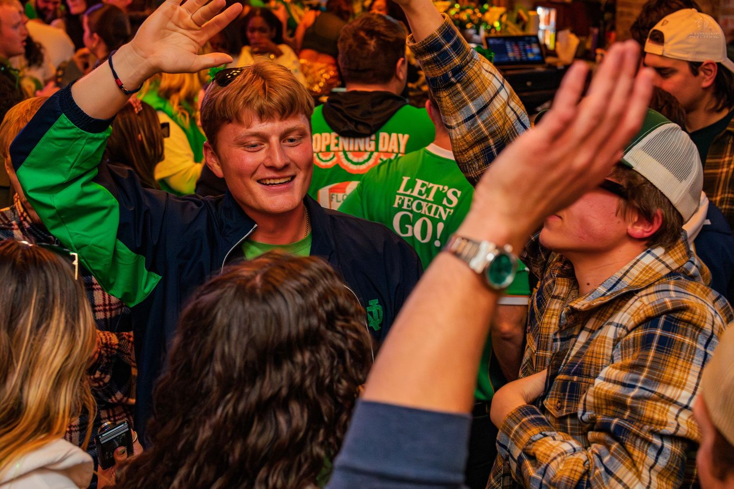 Excited crowd of people celebrating St. Patrick's Day at a packed bar, raising hands.