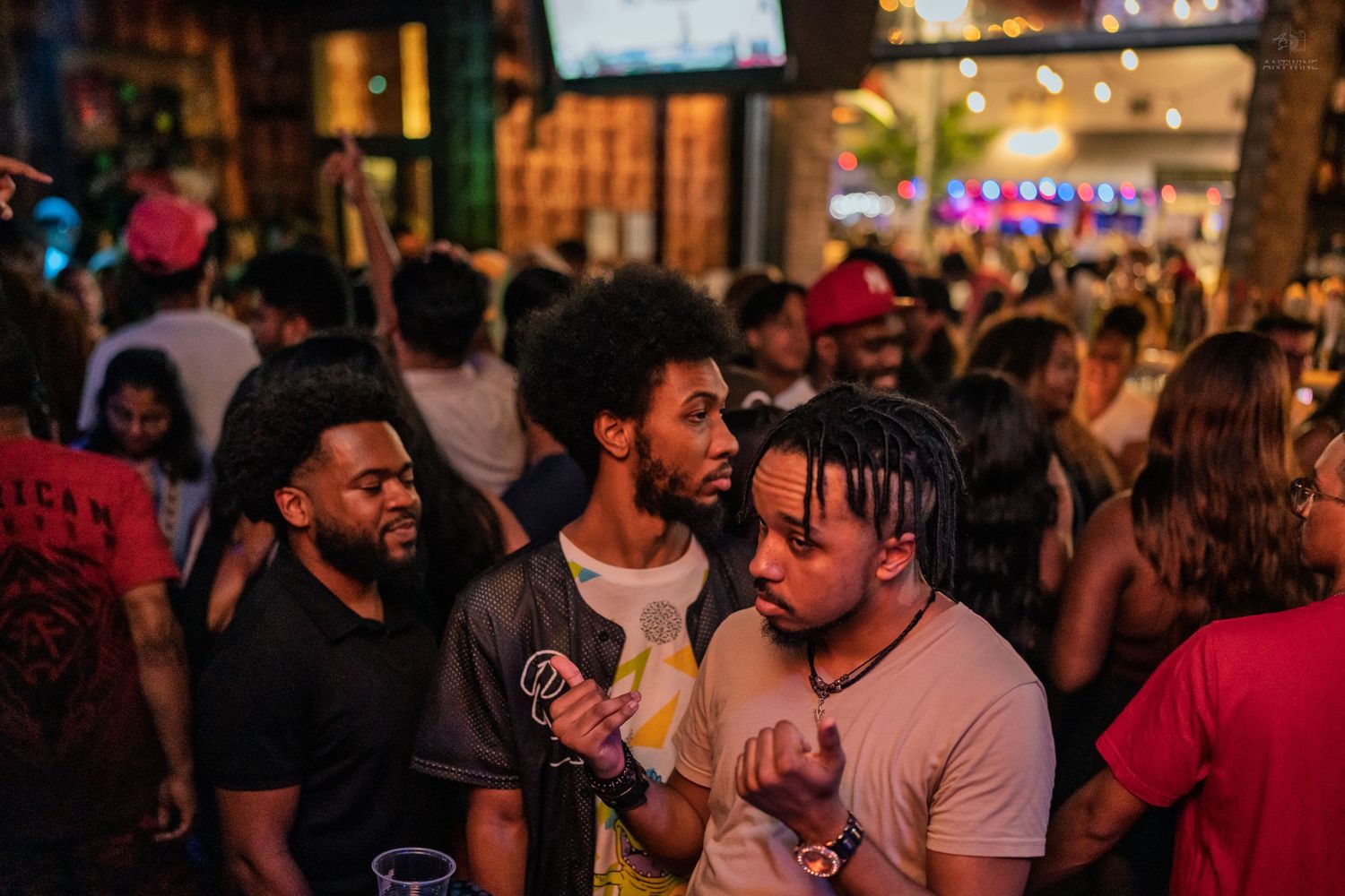 Three young men conversing outdoors at a busy nightlife venue with string lights.