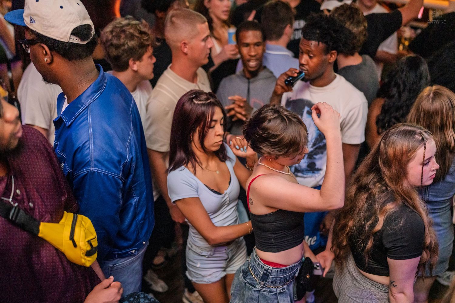 Crowded nightclub dance floor with diverse young adults dancing and socializing in dim lighting.