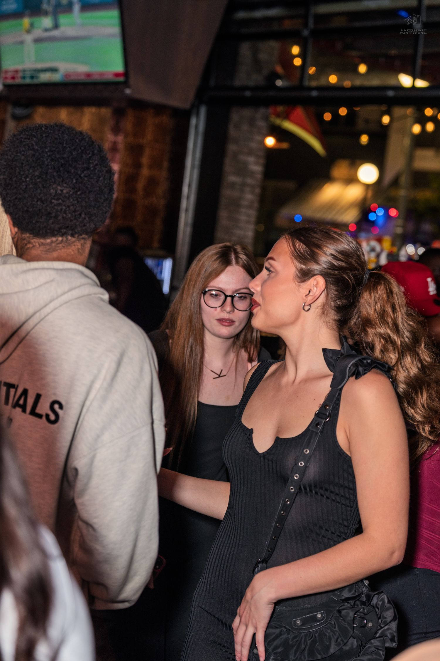 Two women in black outfits socialize at a busy bar while a man in Essentials hoodie stands nearby.