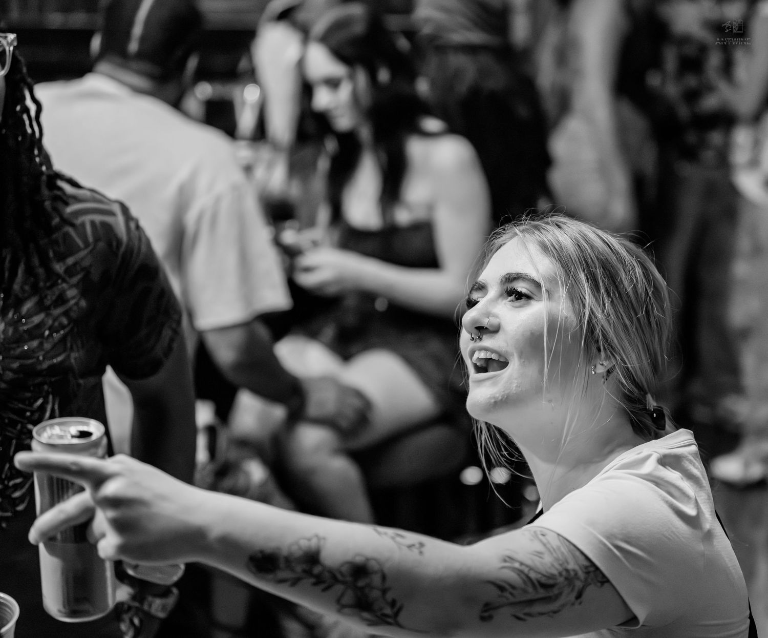 Tattooed woman smiling and holding a beer can at a lively black and white party scene.