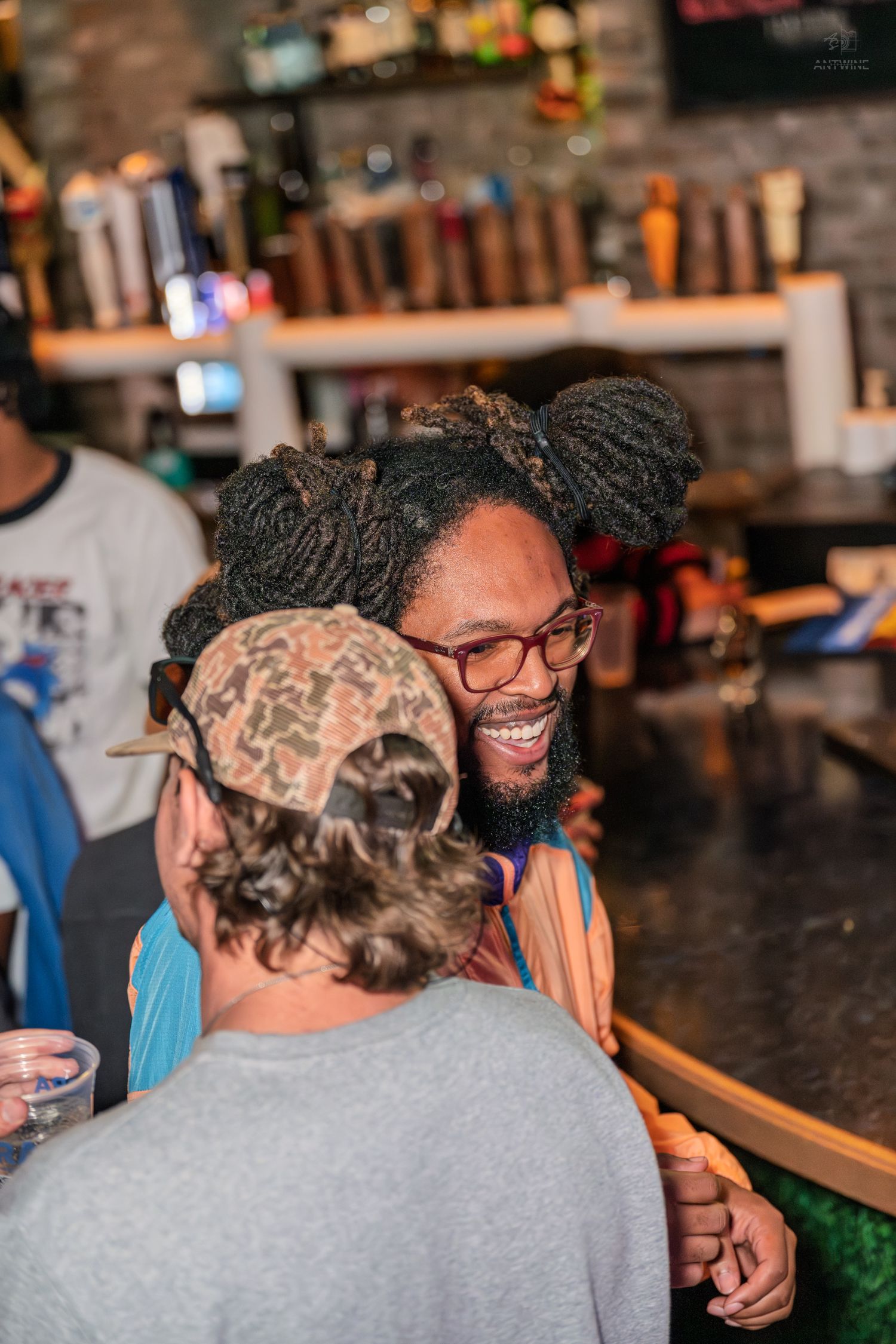 Laughing man with dreadlocks and glasses socializing with friends at a bar counter.