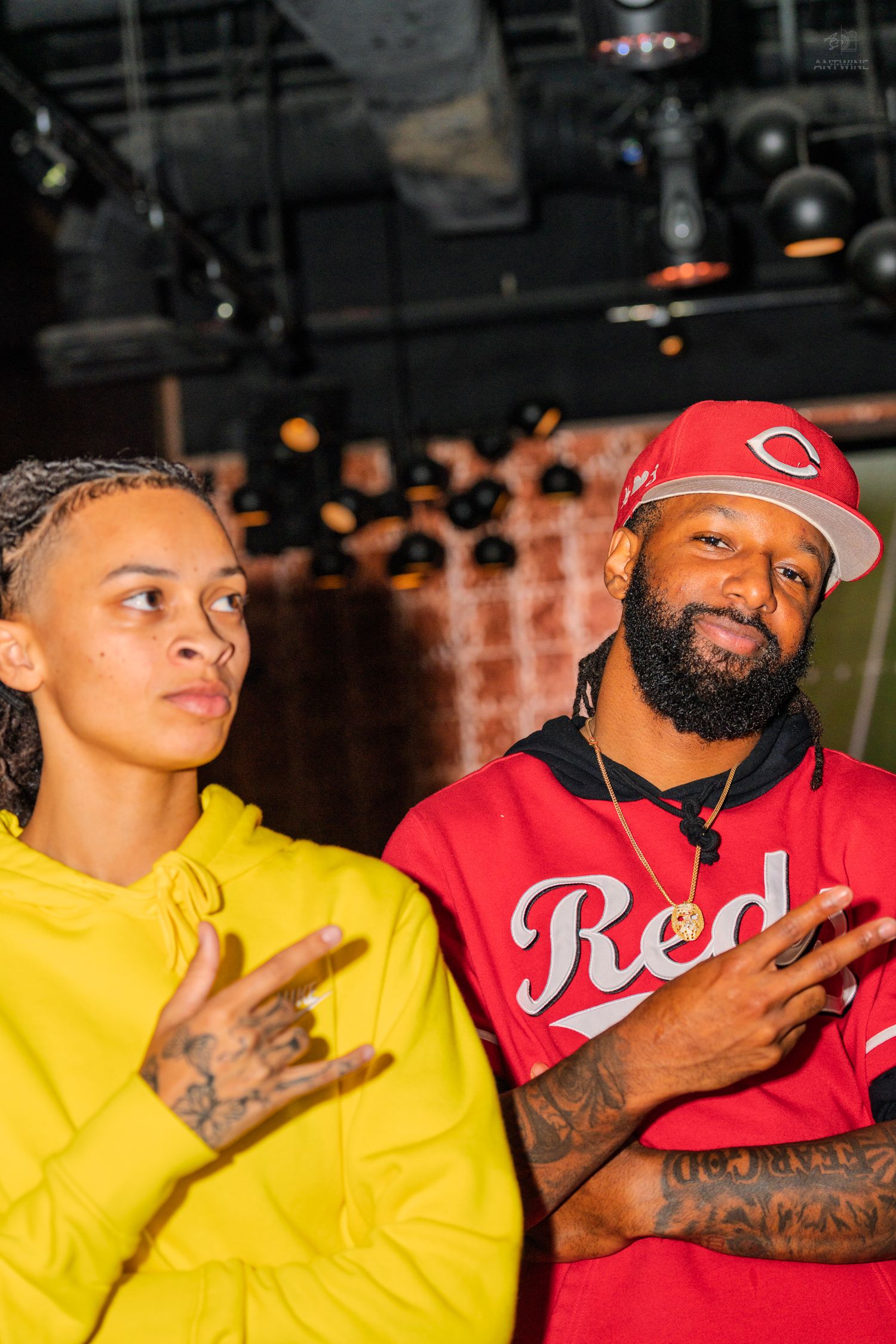 Two people posing at a music venue, one in yellow Nike hoodie and one in red Reds jersey.