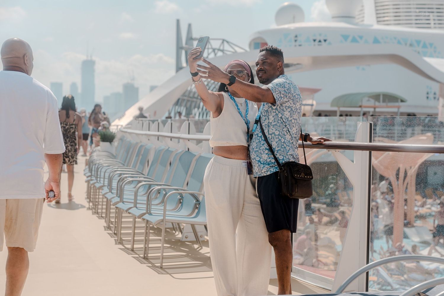 Couple taking a selfie on a sunny cruise ship deck with city skyline visible in the background.
