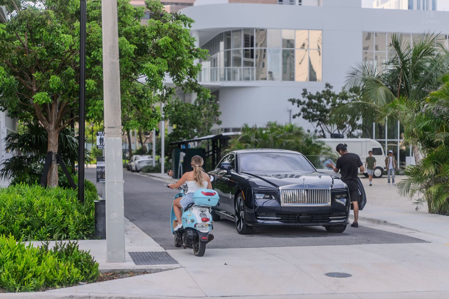 Person riding a scooter past a black Rolls-Royce luxury car outside a modern white building.