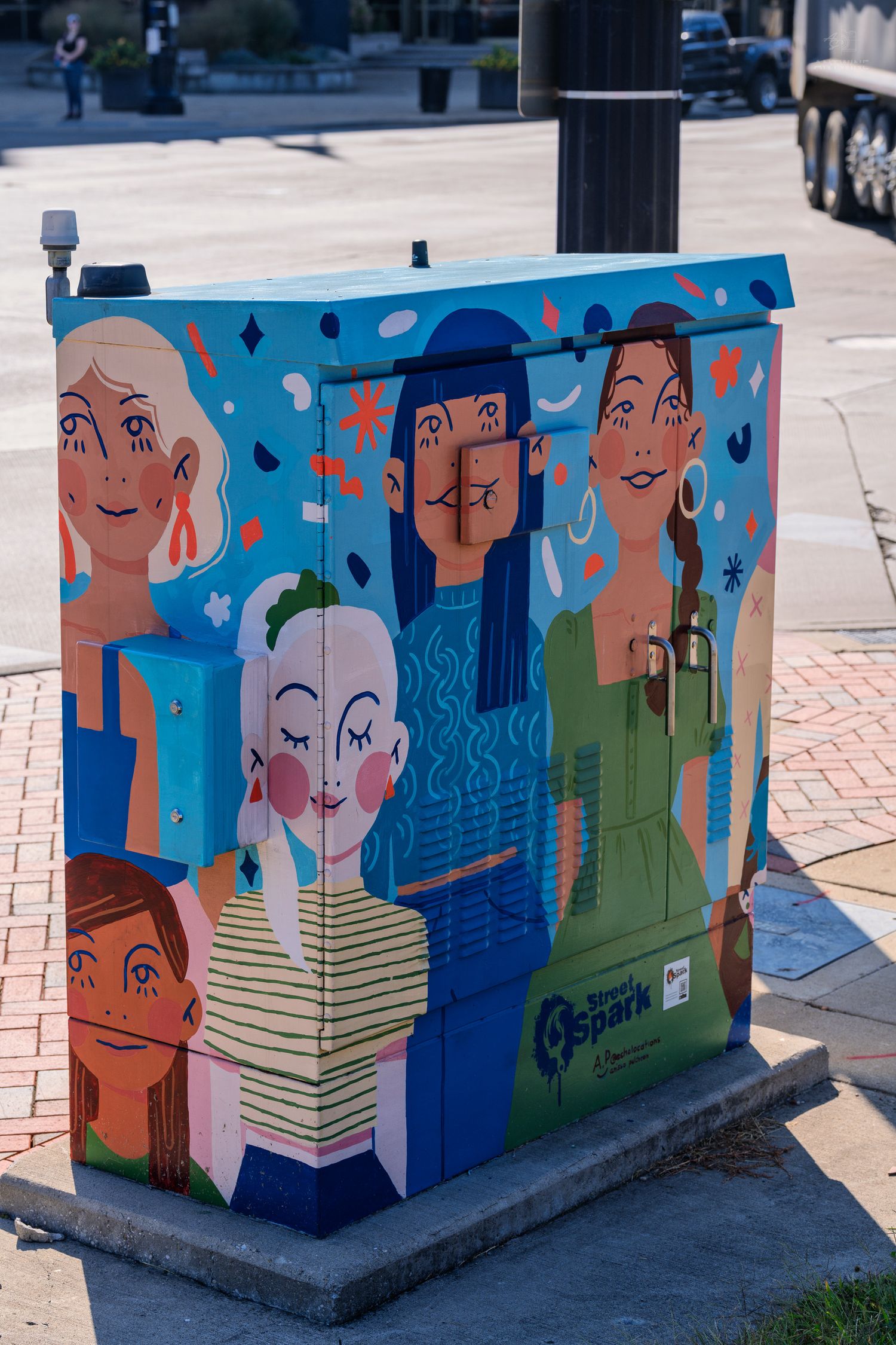 Colorful street art mural on utility box featuring diverse women and children painted in vibrant blues, greens, and pinks.
