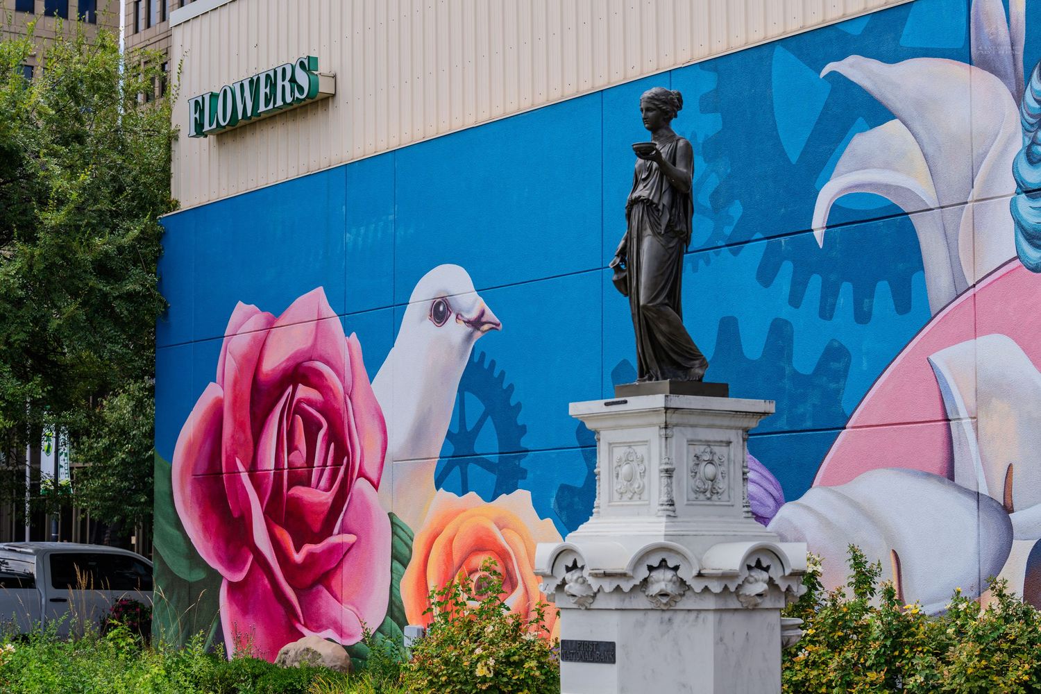 Colorful mural with roses and doves behind a classical statue on a pedestal in an urban setting.