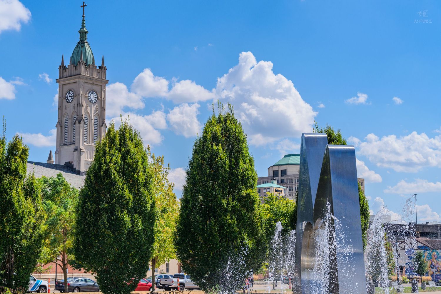 Fountain and trees in a city park with a historic clock tower and blue sky with clouds in the background.