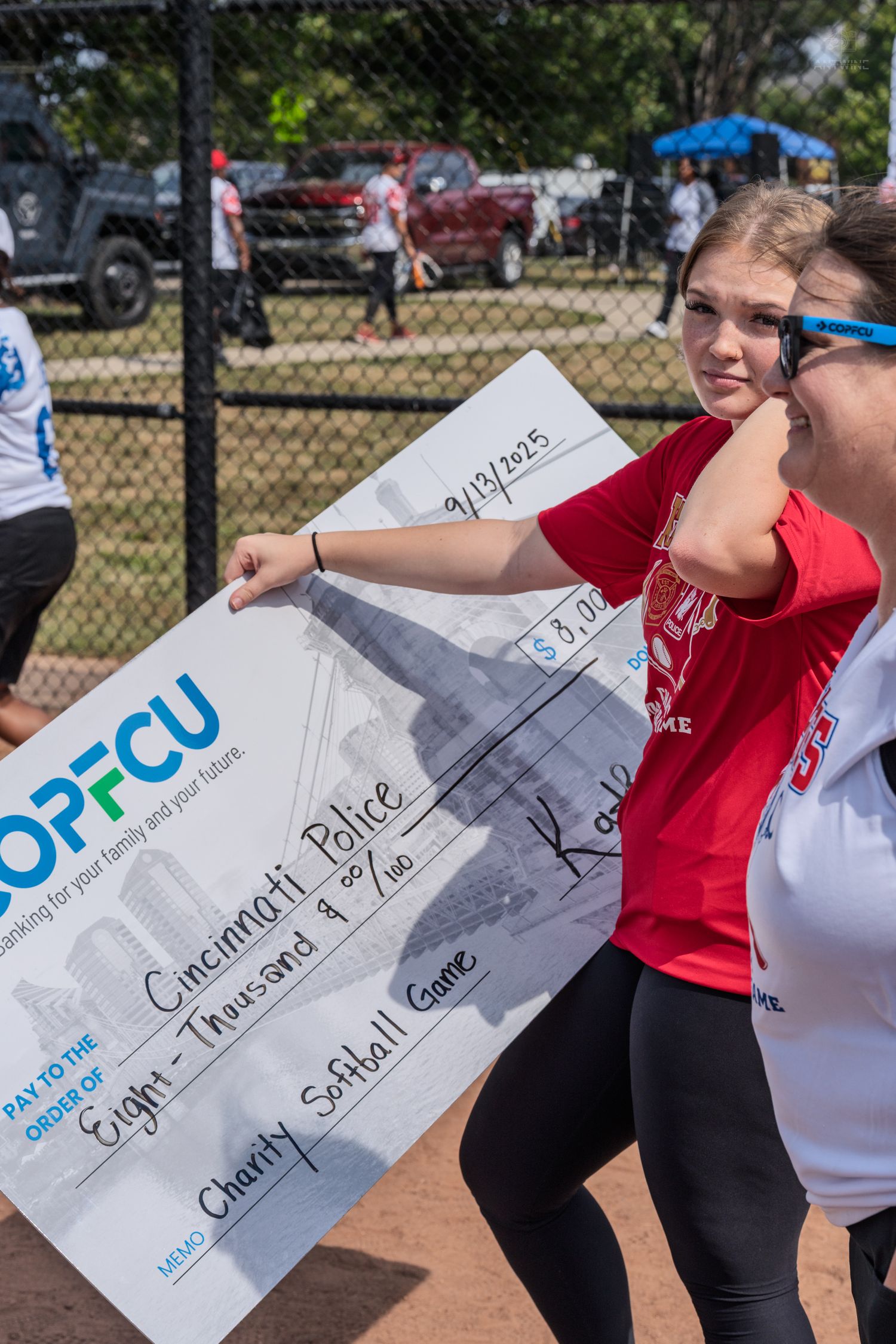 Two girls hold a large OPFCU charity check for $8,000 made out to Cincinnati Police at a charity softball game.