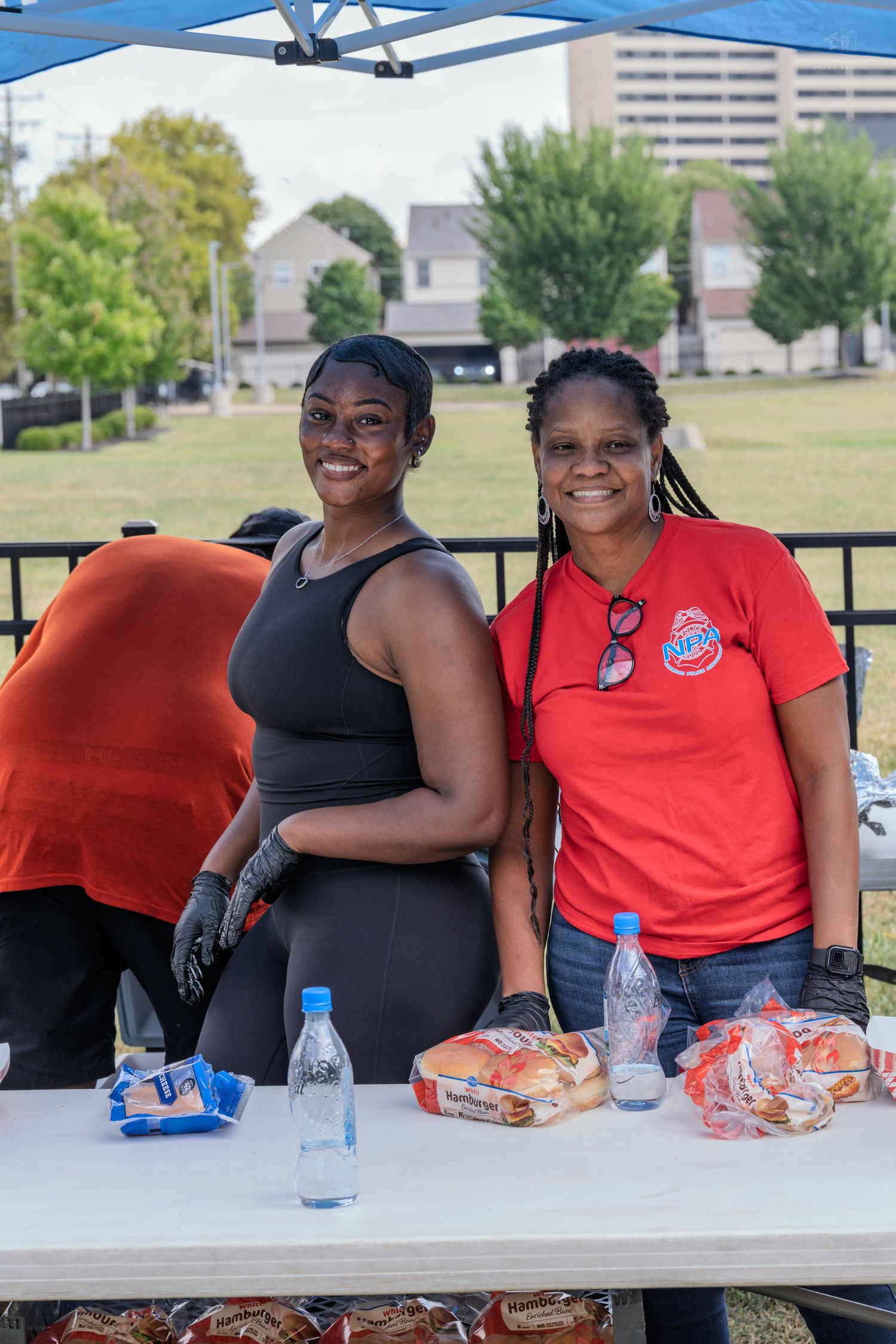 Two women smile at a food service table under a blue tent at an outdoor community event.