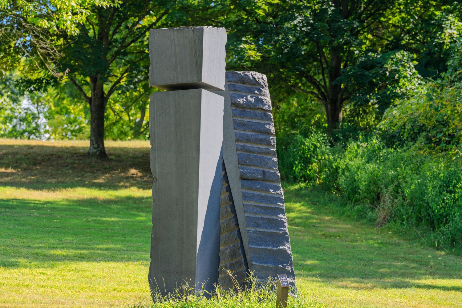 Abstract modern granite sculpture with geometric forms standing on green grass surrounded by trees in an outdoor park setting.