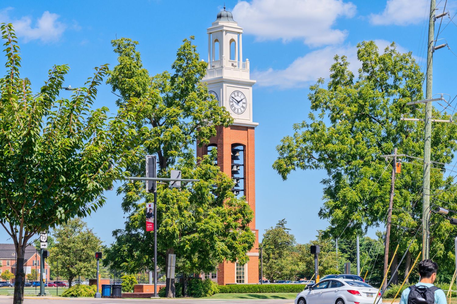 A tall red brick clock tower rises above green trees on a sunny university campus with a clear blue sky.