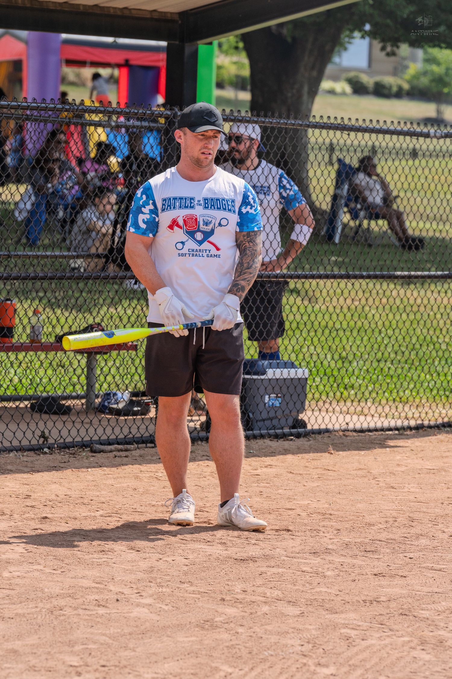 Male player in Battle of the Badges softball jersey holding bat at dugout fence on sunny day.
