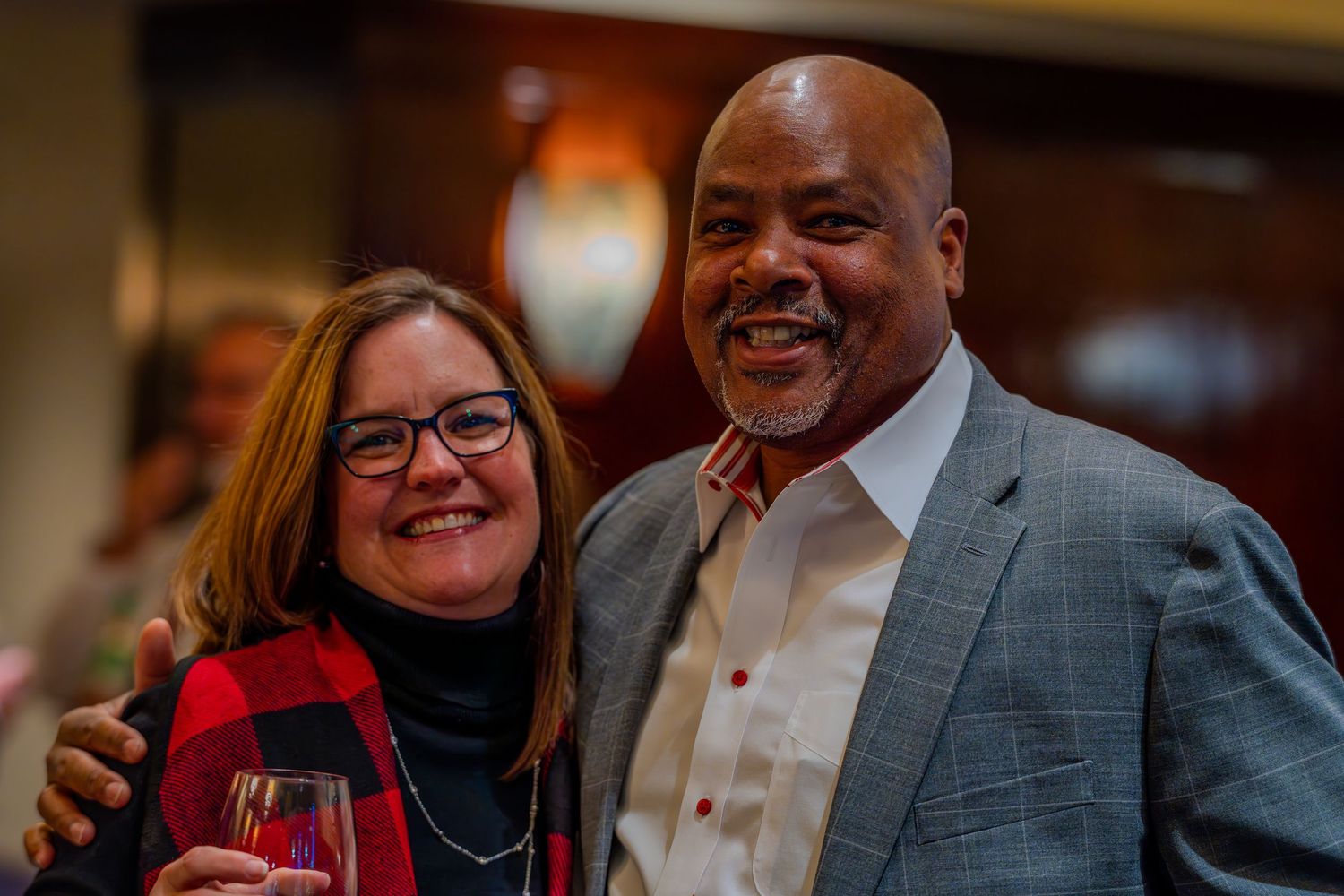 Two smiling people pose together at a social event, woman holding a glass of red wine, wearing festive attire.