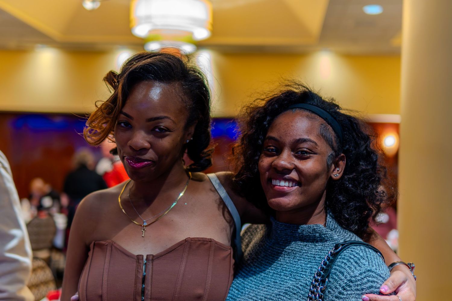 Two young women smiling and posing together at an indoor social event in a warmly lit venue.