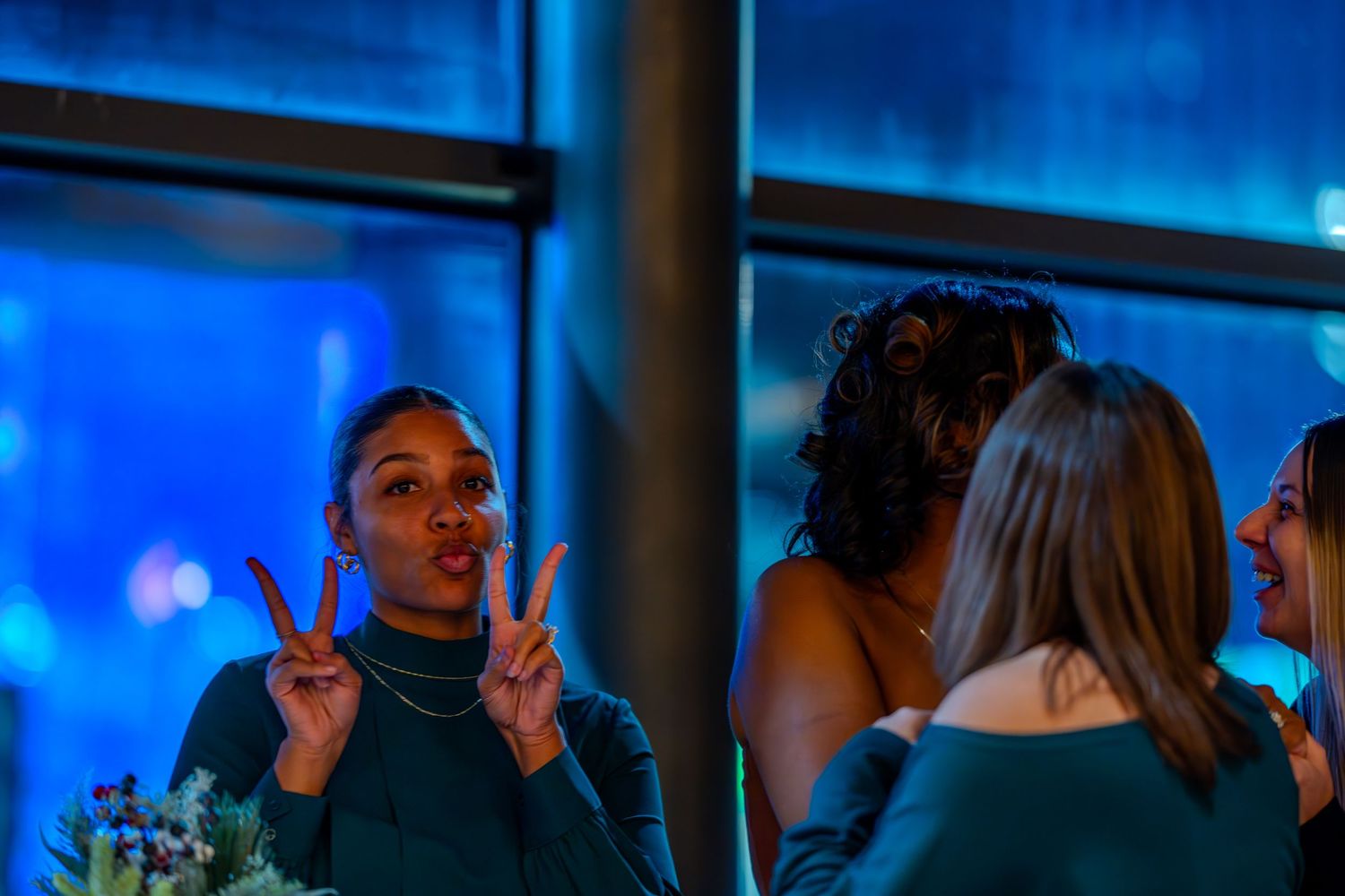 Woman making peace signs with fingers at a blue-lit evening event with friends nearby.