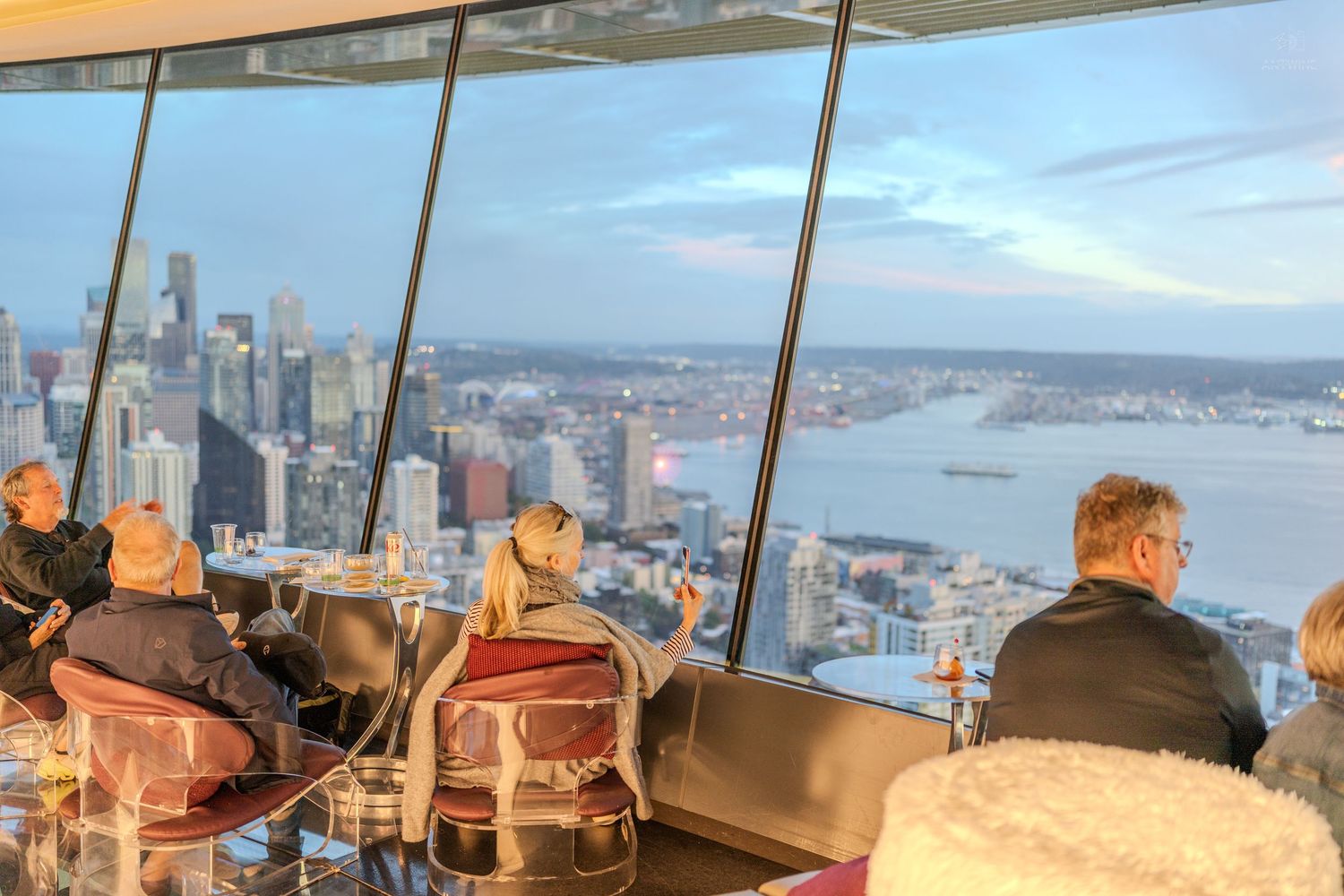 Diners enjoying a panoramic city and waterfront view from a high-rise restaurant at dusk.