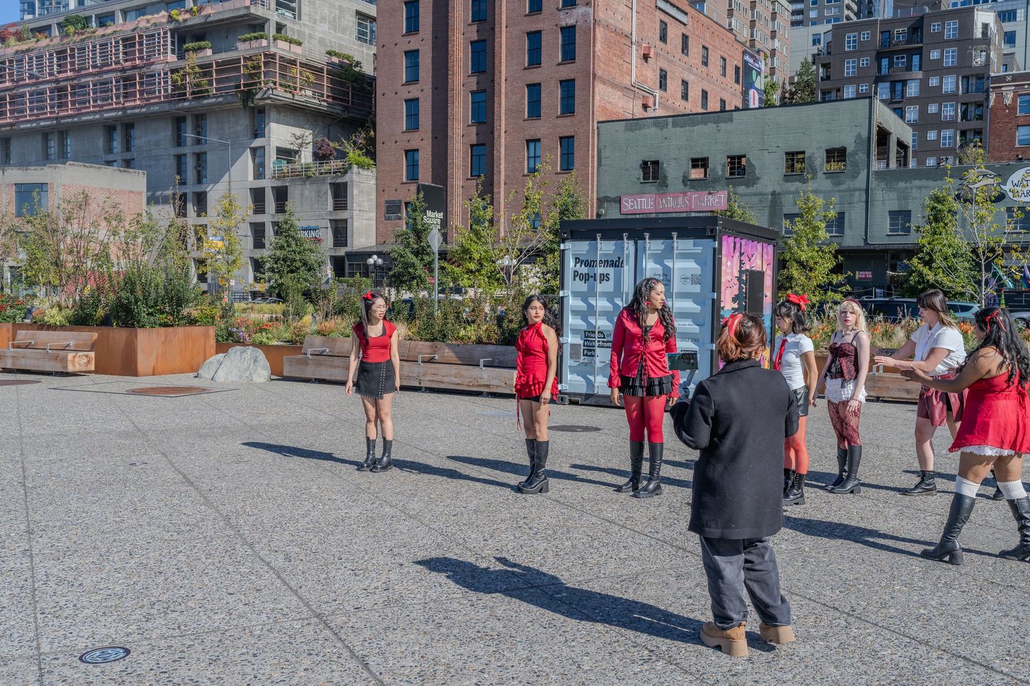 Group of performers dressed in red posing for photographer in sunny Seattle waterfront plaza with urban buildings behind.