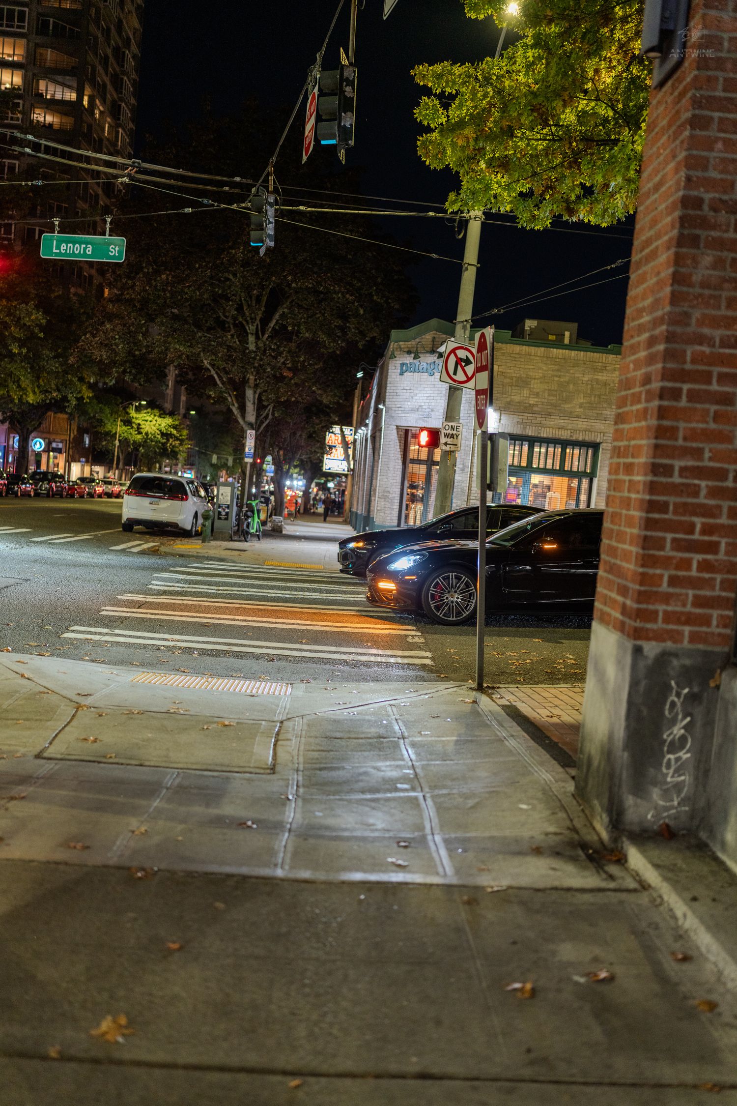 Nighttime urban street corner with crosswalk, parked cars, and illuminated storefronts on a wet sidewalk.
