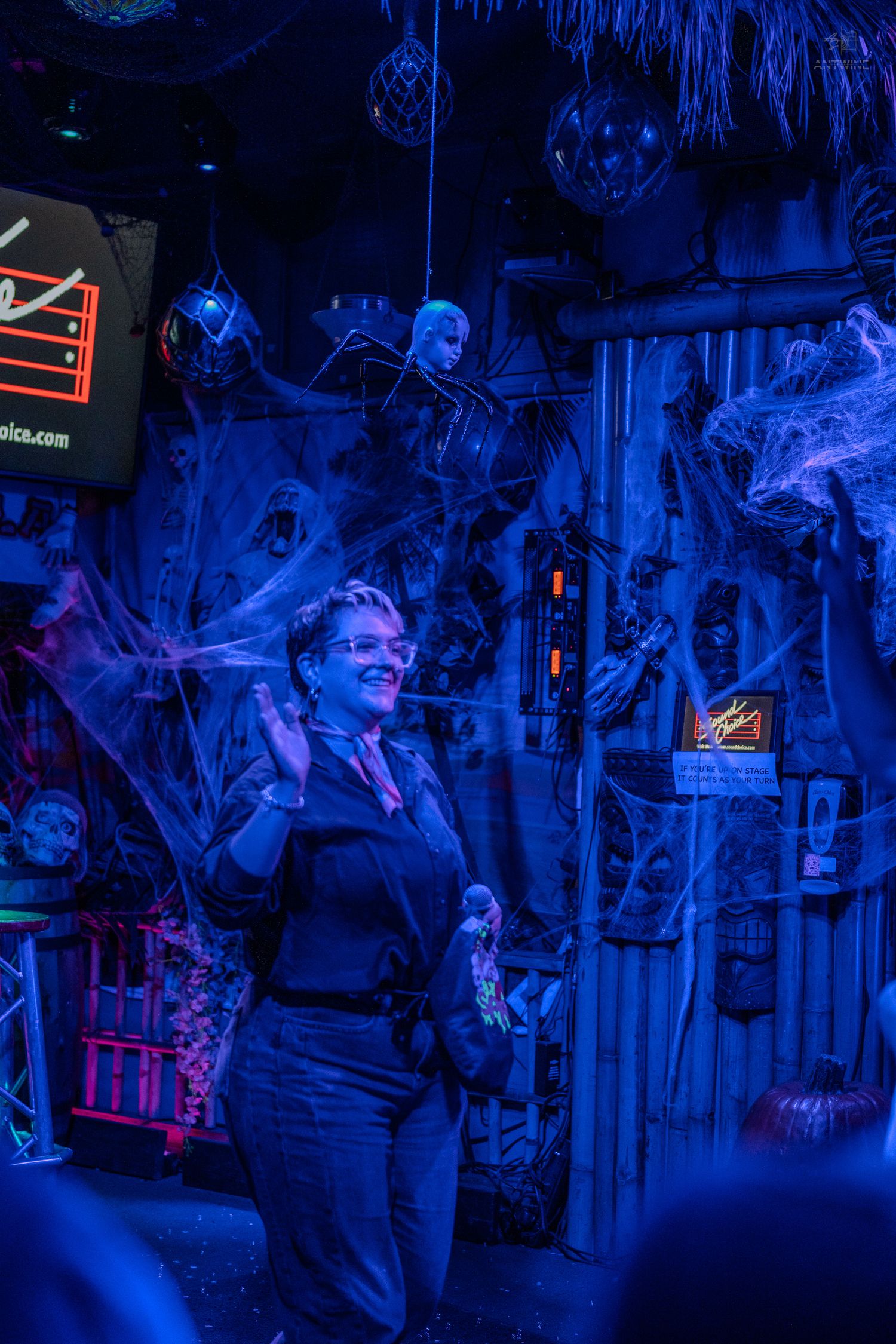 Woman smiling and clapping at a Halloween karaoke bar decorated with spiderwebs under blue neon lights.