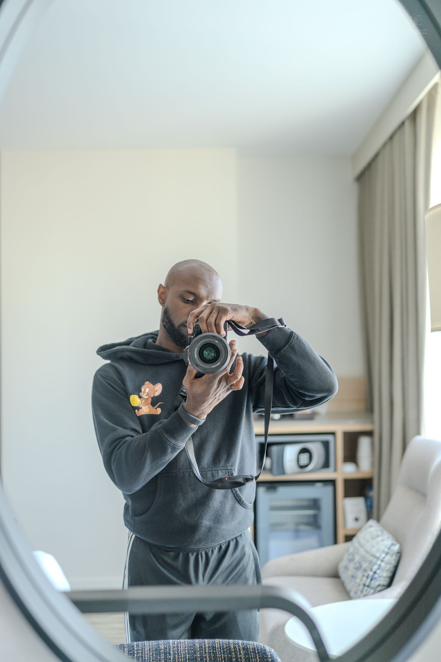 Bald man in gray hoodie takes mirror selfie with professional DSLR camera in bright modern room.