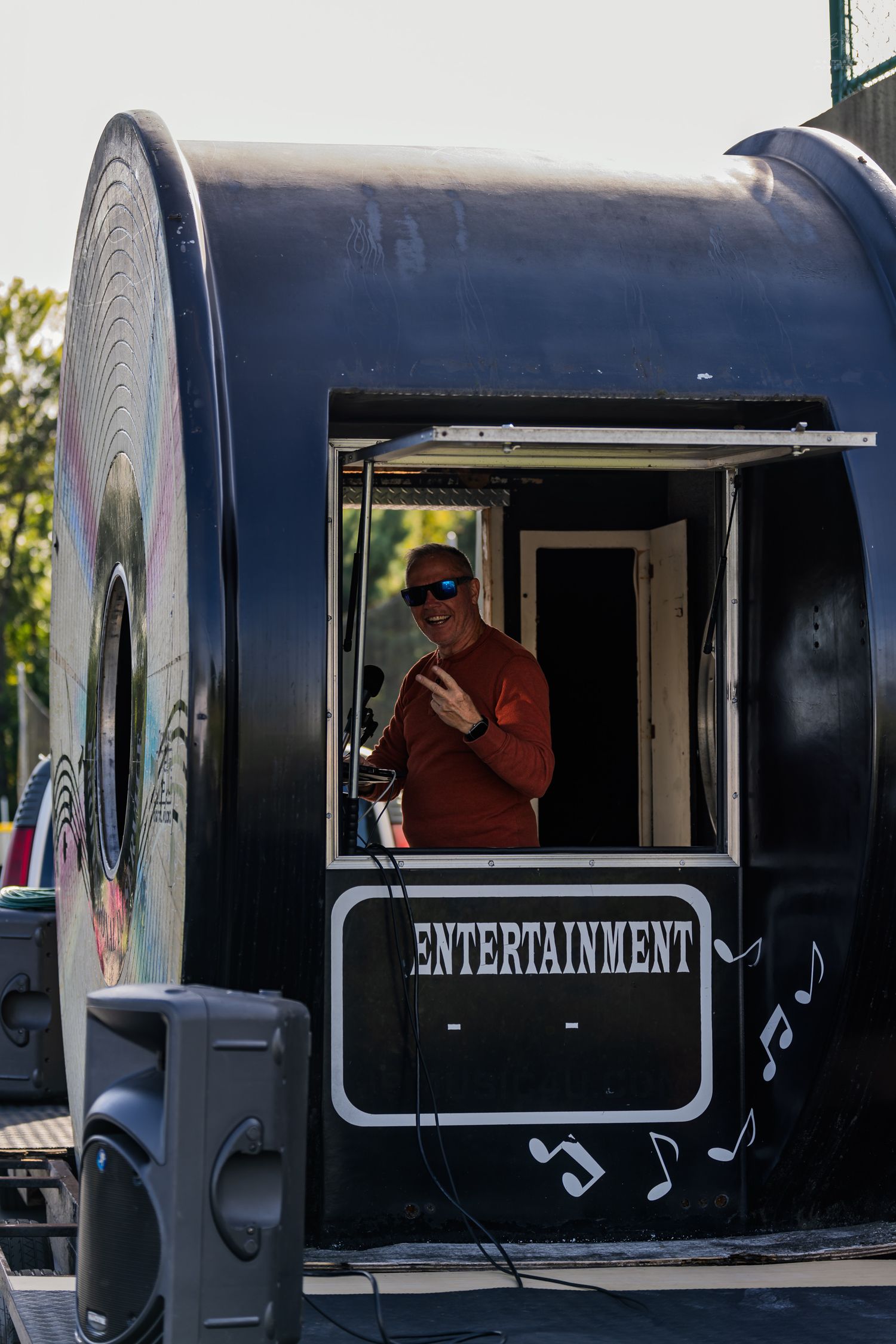 Man in red shirt and sunglasses standing in an entertainment booth, making a hand gesture outdoors.