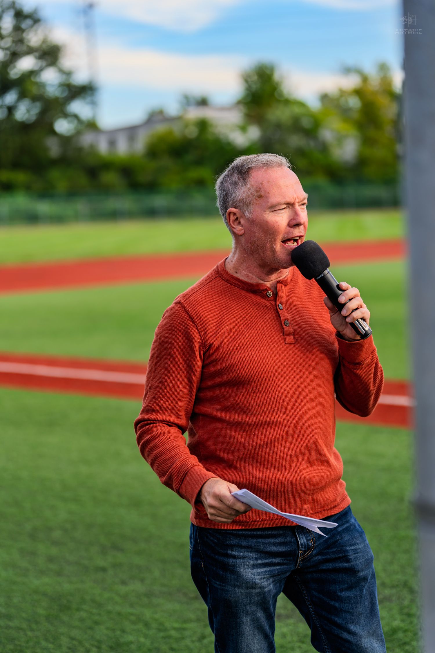 Middle-aged man in red sweater speaking into microphone at outdoor sports field event.