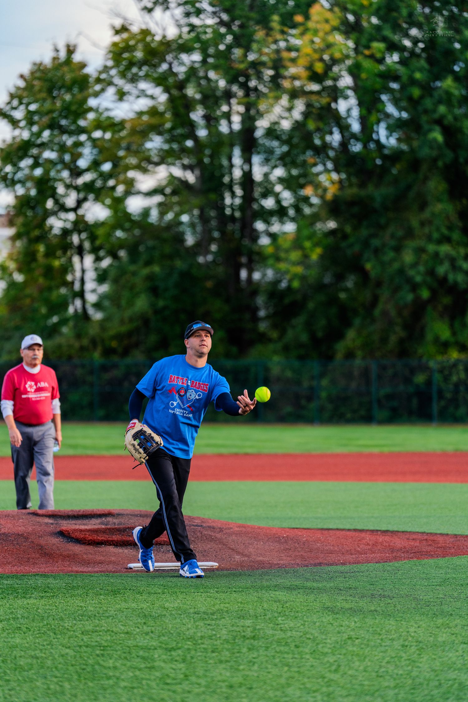 Softball pitcher in blue shirt tosses yellow ball on red turf field during outdoor recreational game.